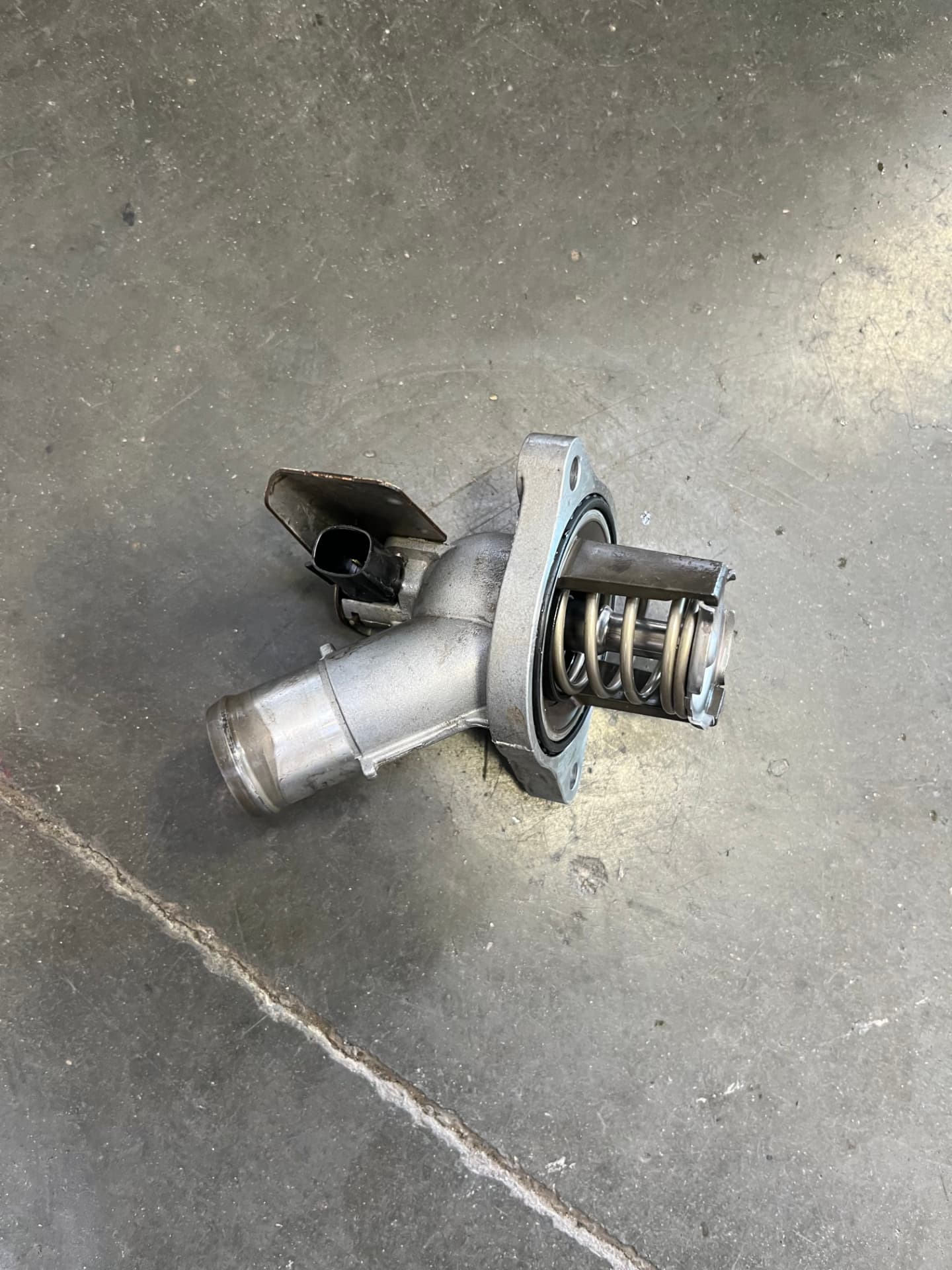 New thermostat housing with its spring and rubber gasket resting on a workshop floor at Johnny's Radiator and Auto Repair